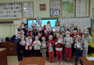Grupa uczniów w czapkach Świętego Mikołaja z nauczycielkami i Świętym Mikołajem. na tle sciany z pomocami edukacyjnymi
